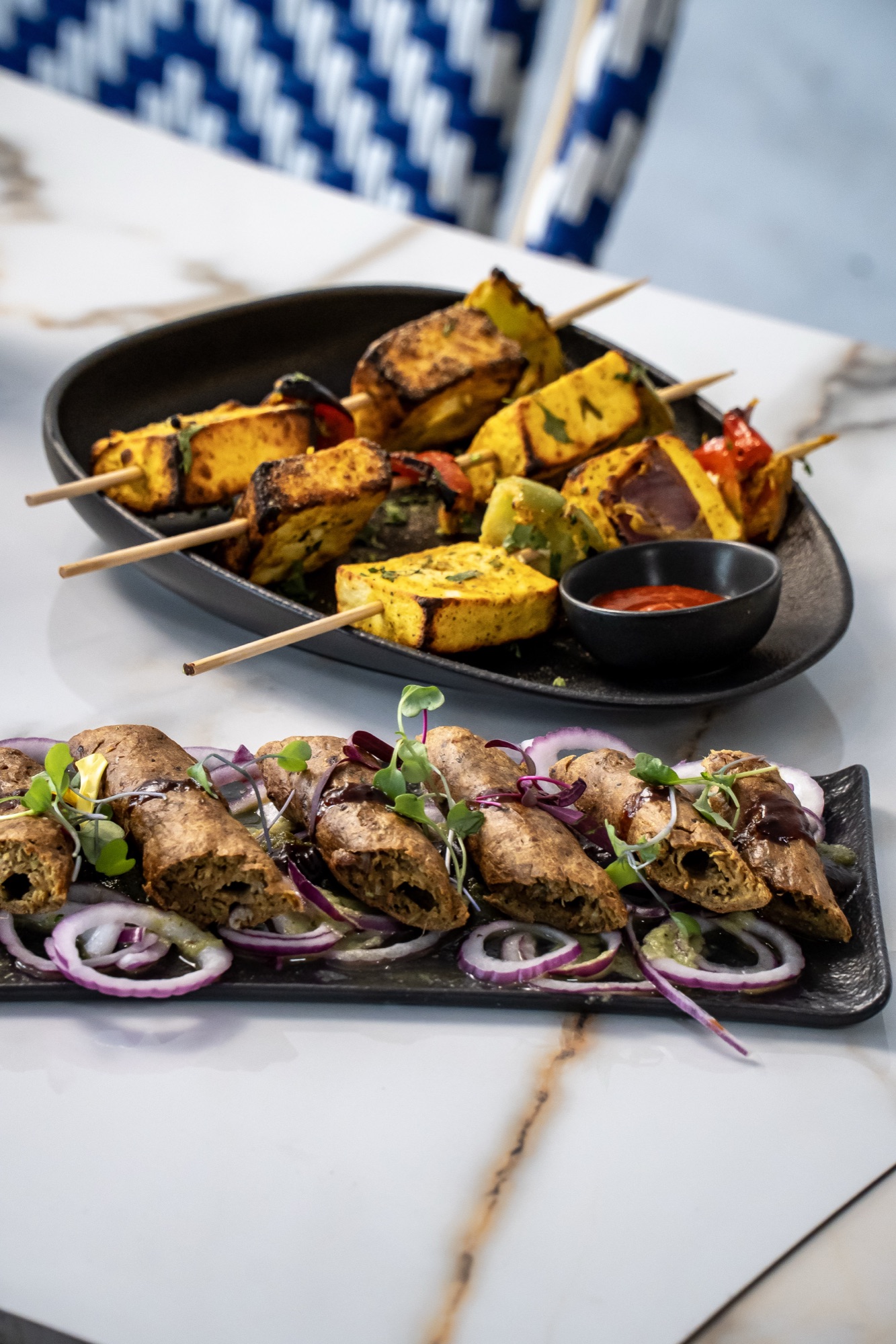 Paneer tikka skewers and seekh kebab platters on marble, blue coastal chairs behind